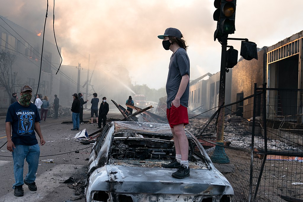 1024px-A_man_stands_on_a_burned_out_car_on_Thursday_morning_as_fires_burn_behind_him_in_the_Lake_St_area_of_Minneapolis,_Minnesota_(49945886467)